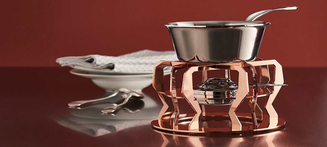Kupfernes Fondueset mit glänzendem Edelstahlkessel, im Hintergrund Teller mit Servietten und Besteck auf einem Tisch vor rotem Hintergrund