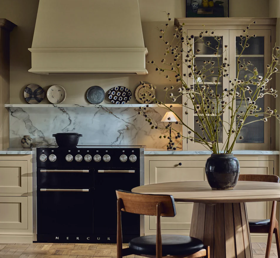 Elegante Küche mit schwarzem Mercury Range Cooker, cremefarbenen Fronten, Marmor-Spritzschutz und rundem Holztisch mit Vase – hochwertiger Design-Gasherd für stilvolle Kochbereiche