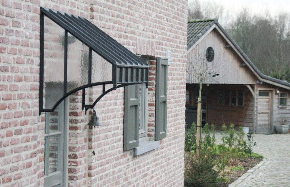 Hochwertiges Vordach aus Glas und Metall für Haustüren – Witterungsschutz im Landhausstil Vordach aus Glas und Metall an einer Hauswand mit Backsteinfassade und Fensterläden, im Hintergrund steht ein Holzhaus