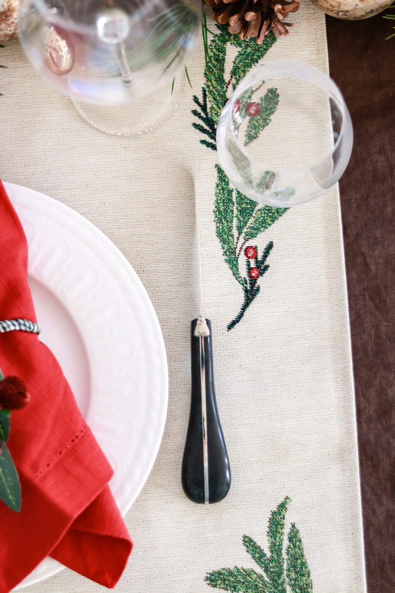 Festlich gedeckter Tisch mit weißem Teller, roter Serviette und Besteck auf besticktem Tischläufer Nahaufnahme eines festlich gedeckten Tisches mit weißem Teller, roter Stoffserviette, schwarzem Besteck und weihnachtlich besticktem Tischläufer mit grünem Zweig und roten Beeren