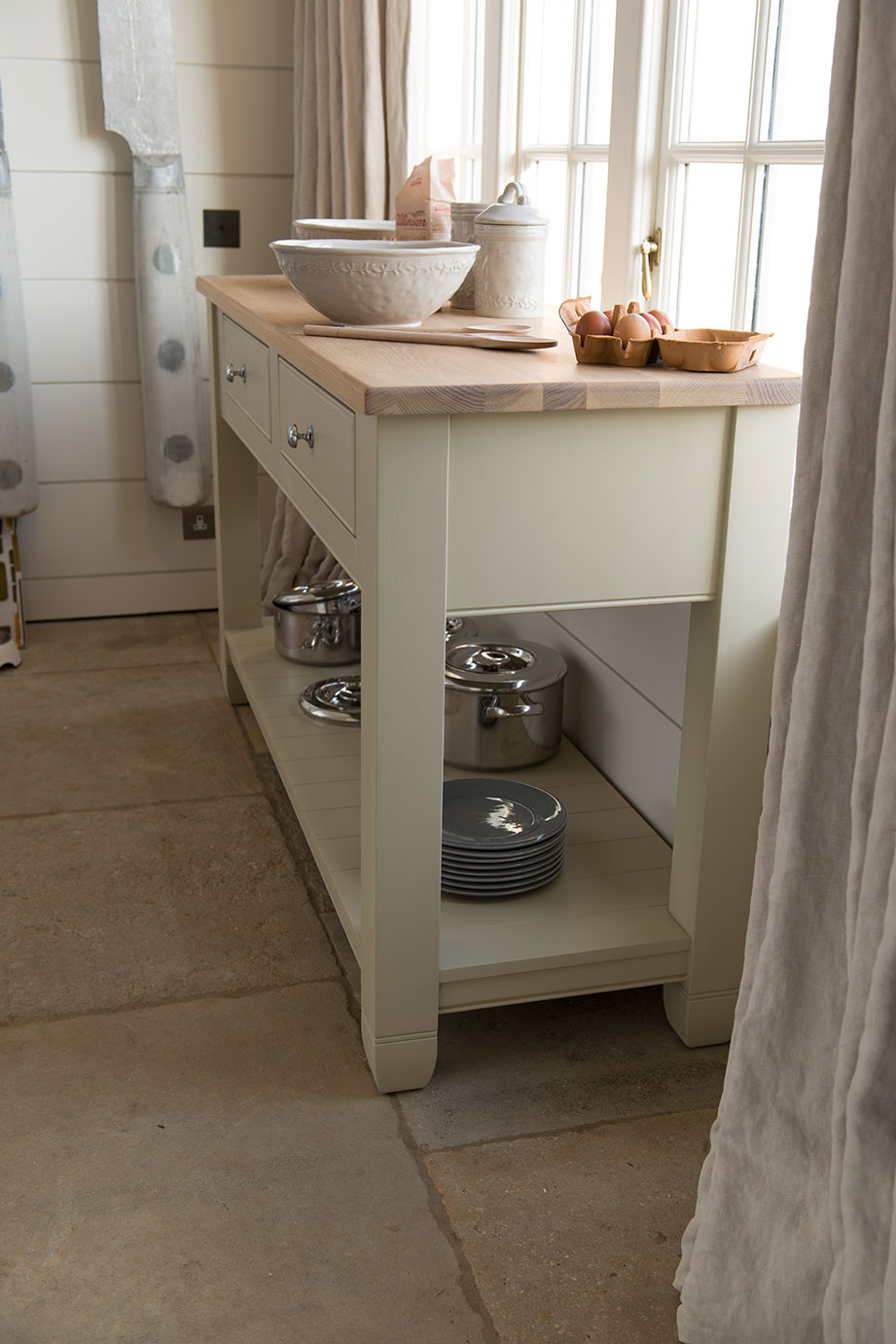 Kücheninsel im Landhausstil aus Holz mit Schubladen und Ablagefläche, dekoriert mit Backzubehör und Geschirr Helle Kücheninsel aus Holz mit Schubladen, Töpfen und Tellern im unteren Regal, auf der Arbeitsfläche Eier, Zucker, Schalen und Kochlöffel