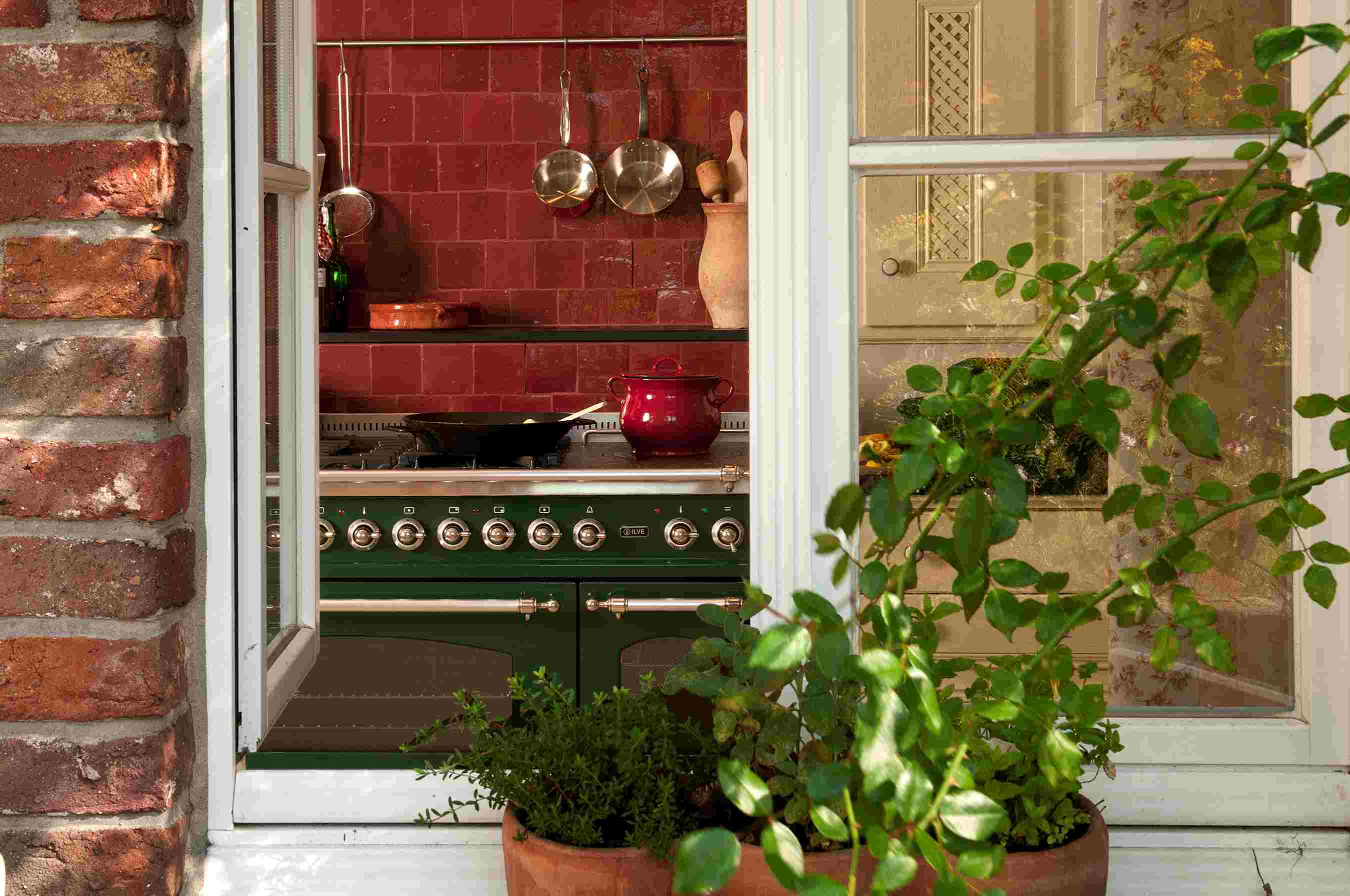 Offenes Fenster mit Blick in eine gemütliche Küche mit Herd, Töpfen und rot gefliester Wand, umgeben von Kletterpflanzen
