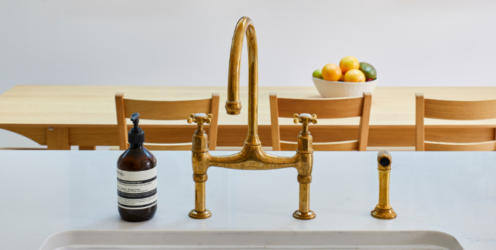 Elegante Küchenarmatur aus Messing mit Seifenspender auf weißer Arbeitsplatte Messingarmatur mit Seifenspender auf einer weißen Küchenarbeitsplatte, im Hintergrund Holztisch mit Stühlen und Schale mit Obst