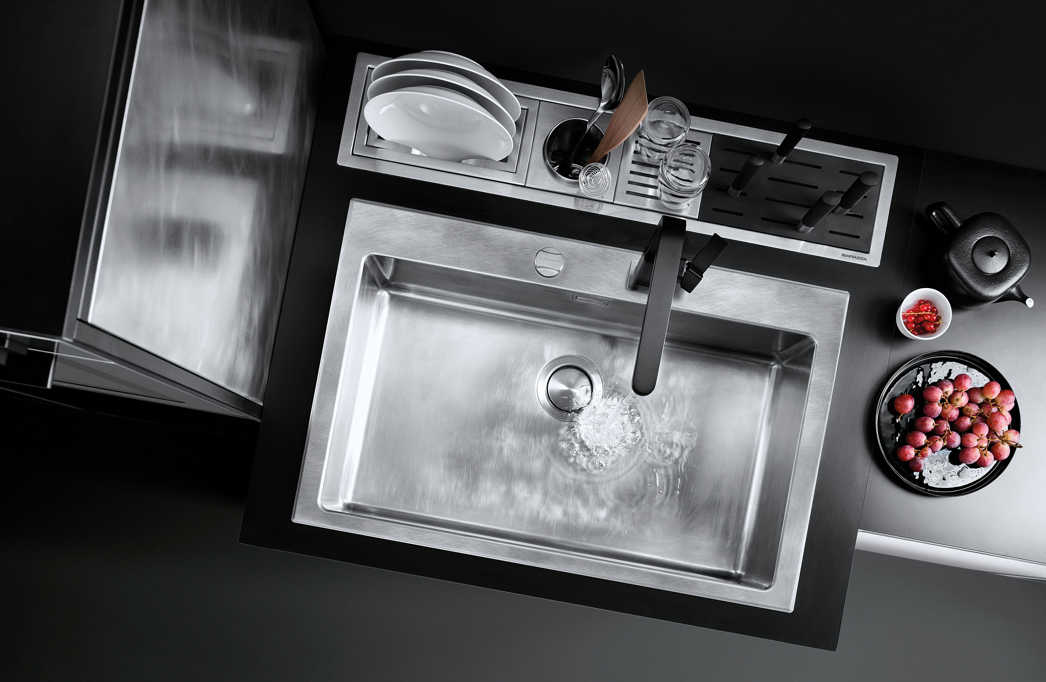 Moderne Edelstahl-Küchenspüle mit schwarzer Armatur und integriertem Abtropfbereich, dekoriert mit Obst und Geschirr Obenaufnahme einer modernen Edelstahlspüle mit schwarzer Armatur, integriertem Abtropfbereich, ablaufendem Wasser, weißen Tellern, Gläsern und einem schwarzen Tablett mit Trauben sowie einer kleinen Schale mit roten Beeren