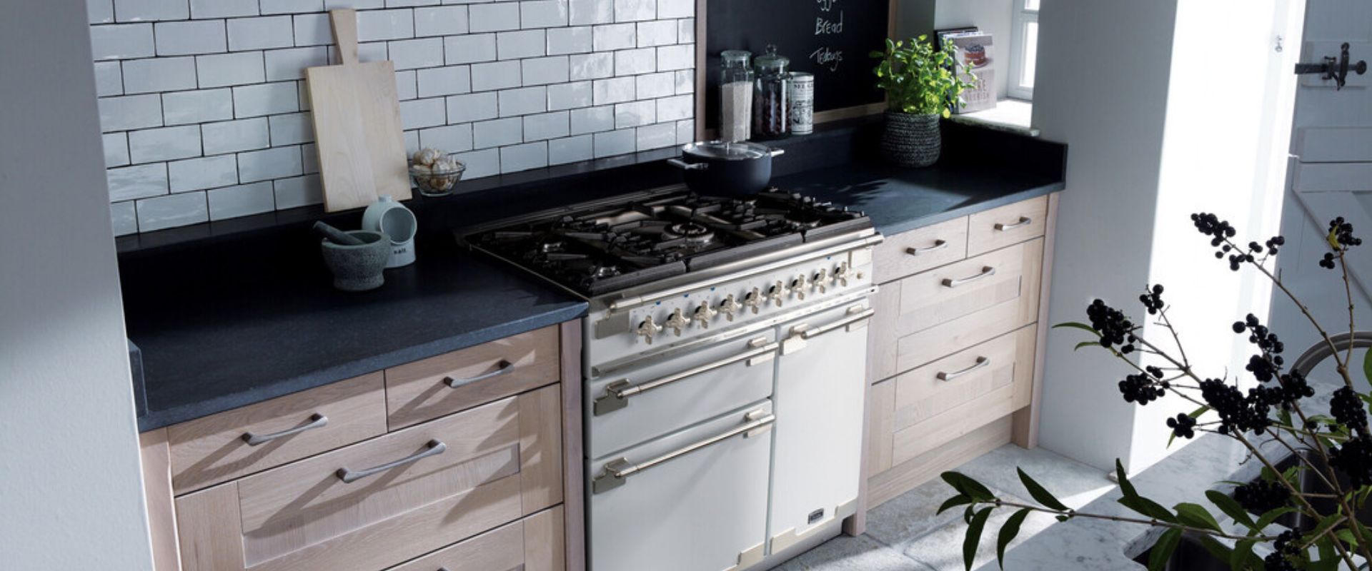 Küchenansicht mit cremefarbenem Standherd und Gaskochfeld, hellen Holzfronten, schwarzer Arbeitsplatte und weißen Metrofliesen – eleganter Range Cooker für Landhausküchen