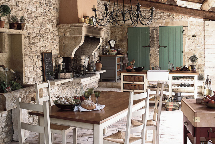 Rustikale Landhausküche mit massivem Holztisch, Steinwänden und Kamin Helle, rustikale Landhausküche mit Steinmauern, großem Holztisch, Kamin, Kronleuchter sowie grünen Fensterläden und Küchenutensilien