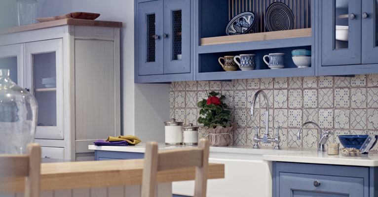 Landhausküche in Blau mit Fliesenspiegel, offenem Regal und Keramikgeschirr Moderne Landhausküche mit blauen Schränken, gemustertem Fliesenspiegel, offenem Regal, Tassen und Tellern sowie Arbeitsfläche mit Spüle und Deko-Pflanze
