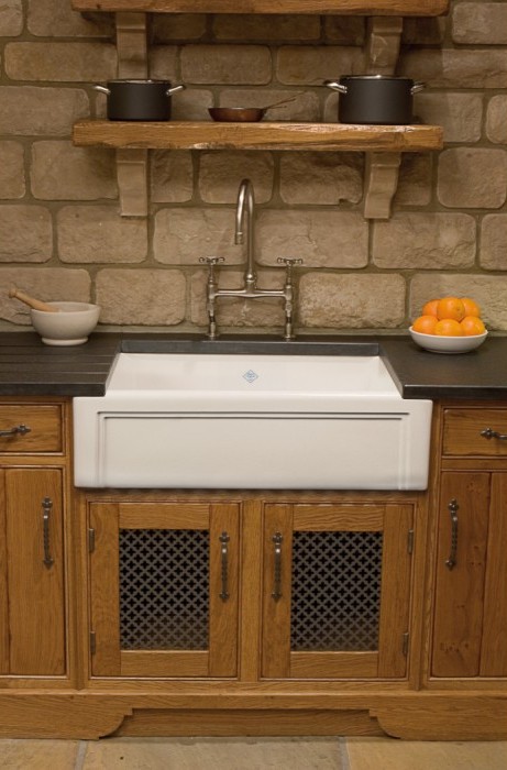 Landhausstil-Küchenspüle aus Keramik mit Holzunterschrank und Wandarmatur Große weiße Keramikspüle im Landhausstil mit Holzunterschrank, Wandarmatur, schwarzer Arbeitsplatte, Regalen mit Töpfen und Schale mit Orangen