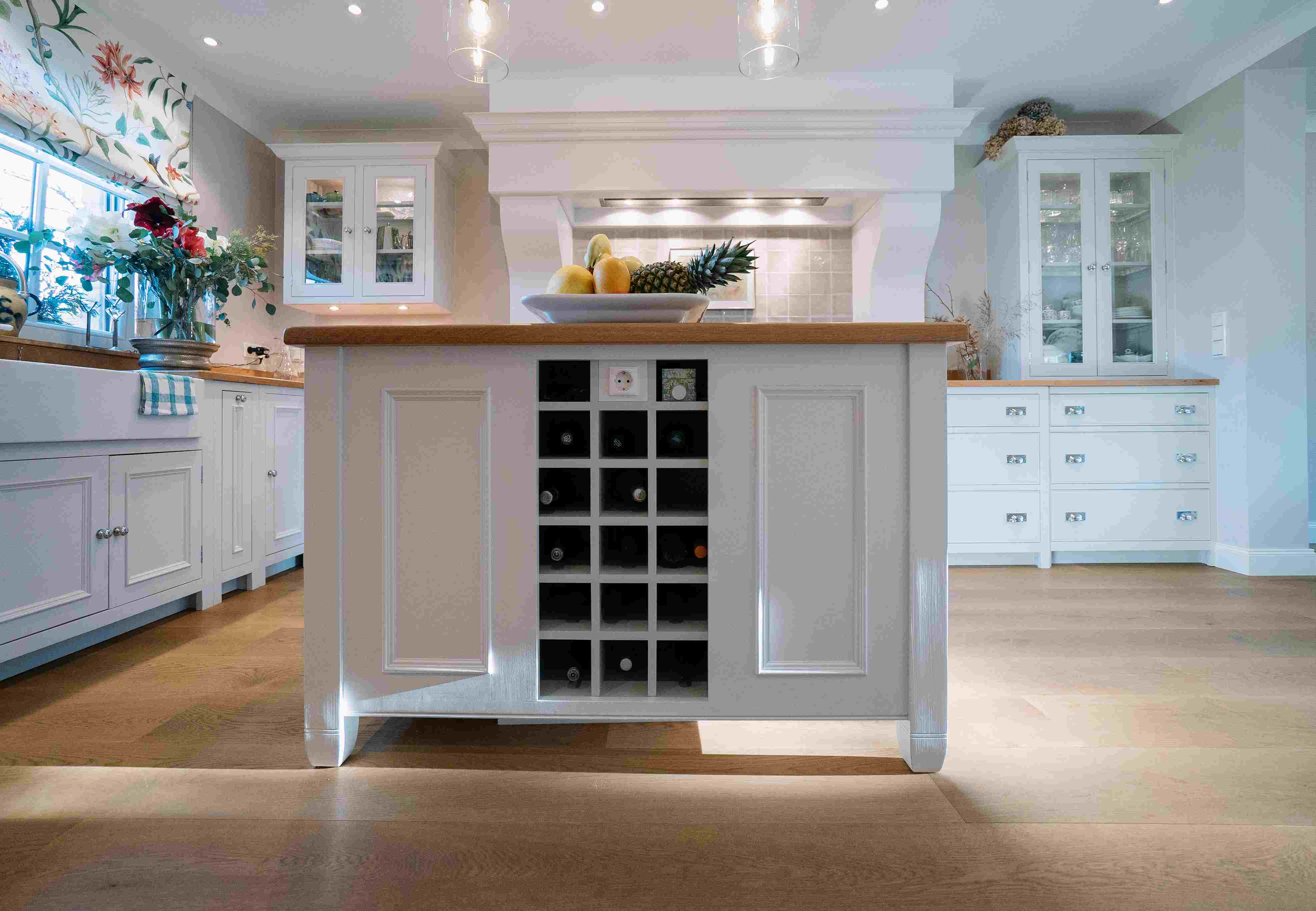 Moderne Landhausküche mit Kochinsel, Weinfächern und hellen Fronten Helle Landhausküche mit Kücheninsel, integriertem Weinregal, Obstschale, Blumen und viel Stauraum