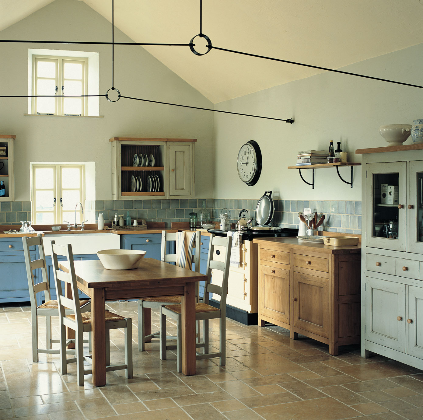 Gemütliche Landhausküche mit Holz-Esstisch, blauen und weißen Schränken sowie großen Fenstern Helle Landhausküche mit Holz-Esstisch, vier Stühlen, blauen und weißen Küchenschränken, Wandregalen und einer Wanduhr