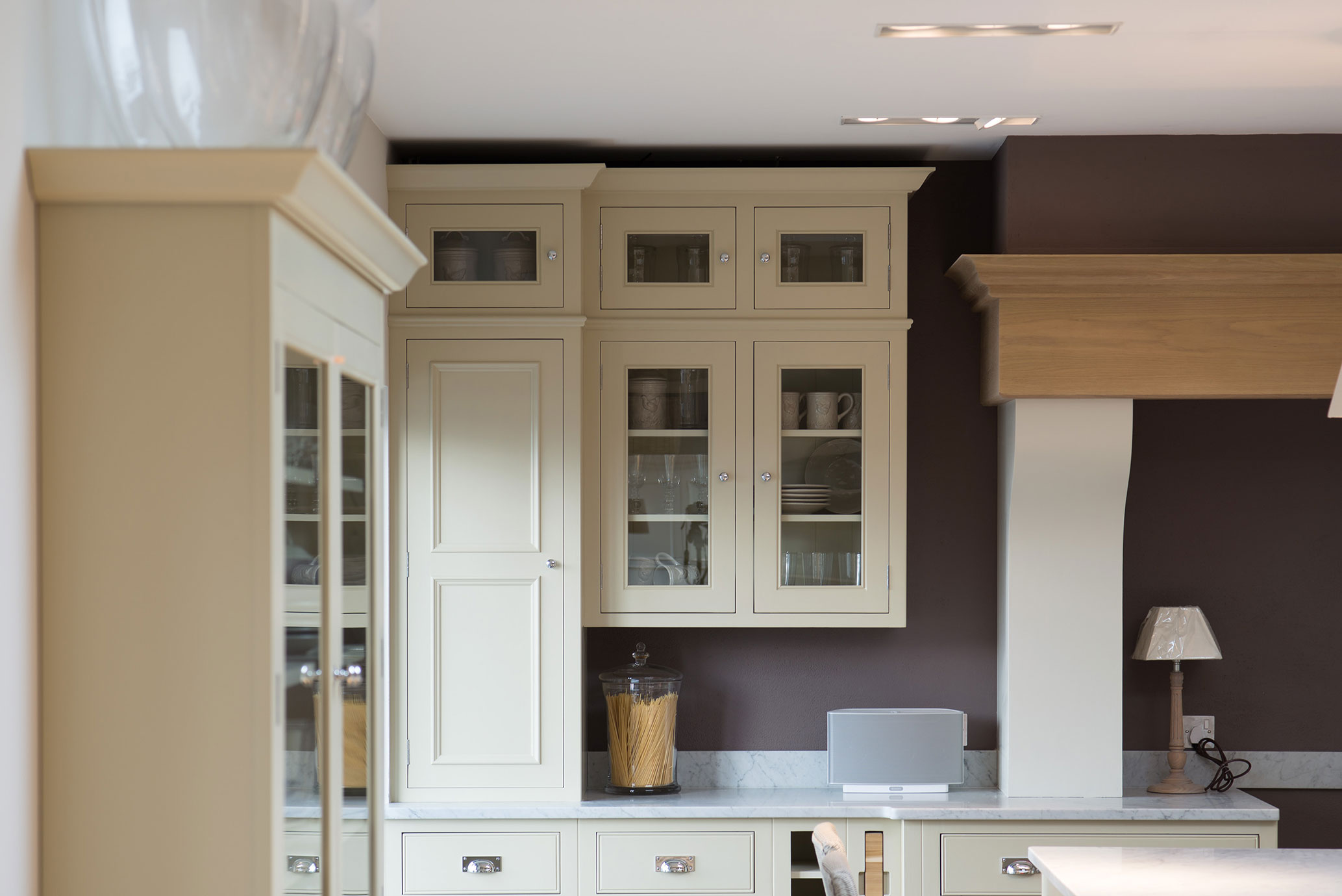 Hochwertige cremefarbene Küchenmöbel mit Glastüren und modernen Details Moderne cremefarbene Küchenschränke mit Glastüren, gefüllt mit Geschirr und Einmachgläsern, auf einer Küchenarbeitsplatte mit Glasbehälter, Lautsprecher und Tischlampe.