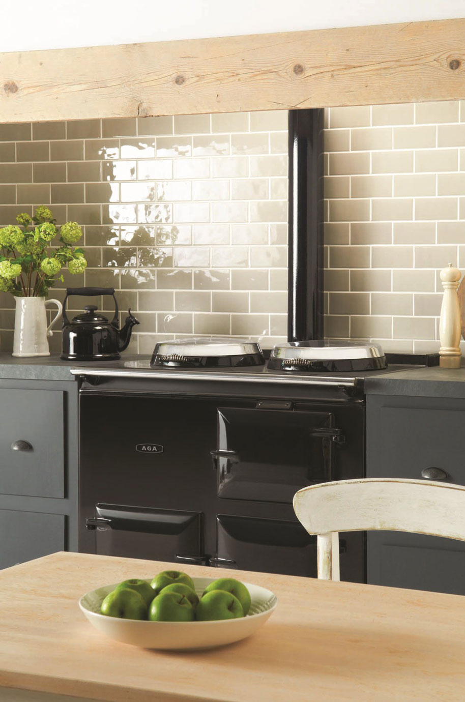 Landhausküche mit schwarzem AGA-Herd, cremefarbenen Metrofliesen und frischen grünen Äpfeln in der Schale Stimmungsvolle Landhausküche mit klassischem schwarzen AGA-Herd, cremefarbenen Wandfliesen, einer Schale grüner Äpfel auf Holztisch und Dekoration wie Teekanne und Vase