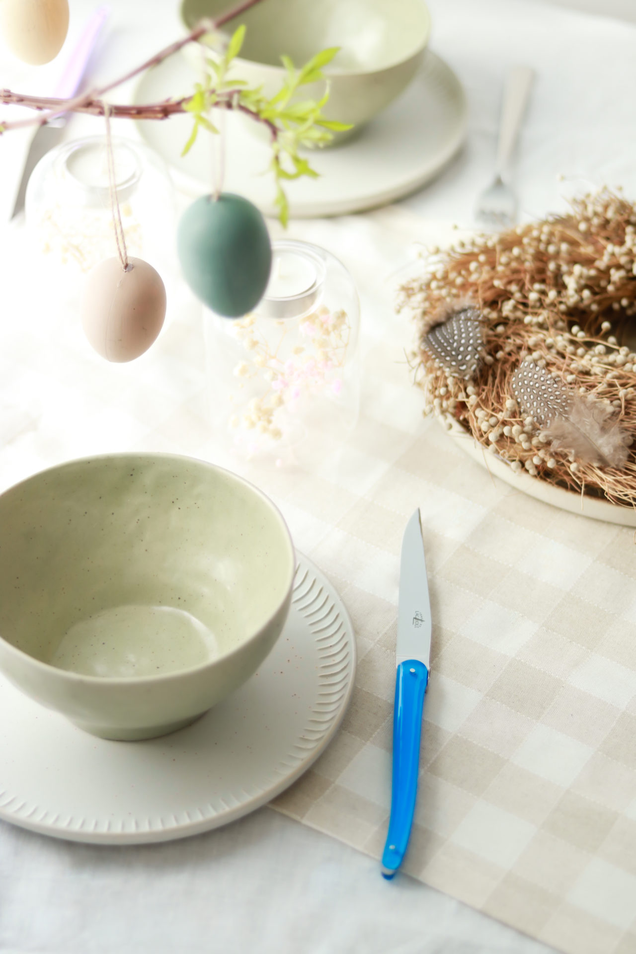 Frühlingshaft gedeckter Tisch mit Keramikschale, Ostereiern und Deko-Kranz Hell gedeckter Tisch mit Keramikschale auf Teller, blauem Messer, Ostereiern an Zweigen und dekorativem Kranz mit Federn