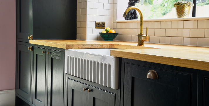 Moderne Landhausküche mit Holzarbeitsplatte, schwarzer Front und Keramikspüle Küchenzeile mit schwarzer Holztür, massiver Holzarbeitsplatte, goldener Armatur und weißer Keramikspüle unter dem Fenster