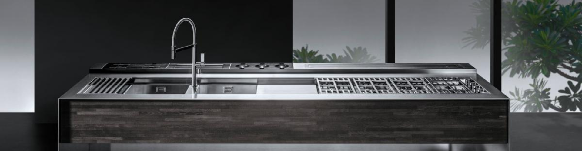 Moderne Kücheninsel mit Edelstahloberfläche, integriertem Kochfeld, Spülbecken und minimalistischer Armatur Elegante, moderne Kücheninsel aus Edelstahl mit Kochfeld, Spüle und dunkler Holzfront, vor großen Fenstern mit grüner Pflanzenkulisse