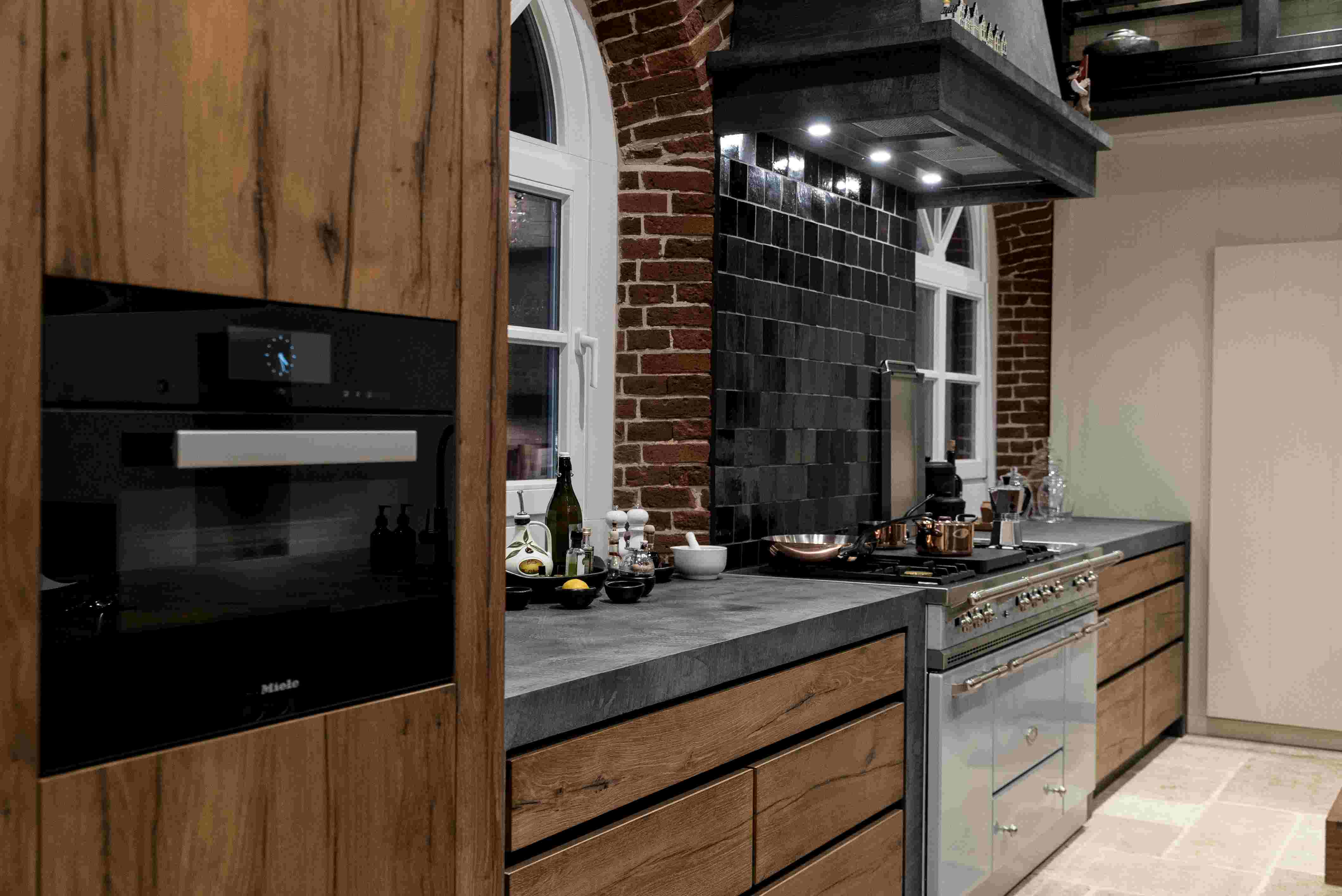 Innenansicht einer modernen Küche mit dunklen Holzschränken, eingebautem Backofen und einem Gasherd mit schwarzen Fliesen an der Wand.