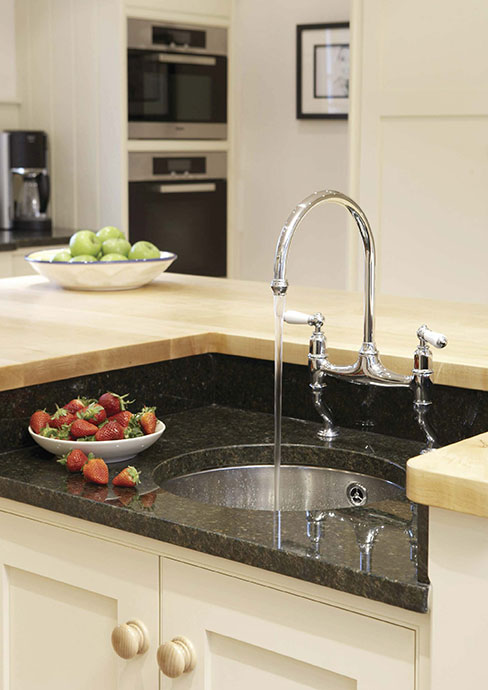 Moderne Küchenarmatur mit fließendem Wasser an Spüle, Obstschale mit frischen Erdbeeren auf Granit-Arbeitsplatte Küchenspüle mit moderner, gebogener Wasserarmatur, daneben eine Schale voll frischer Erdbeeren auf einer schwarzen Granitplatte in einer hellen Küche