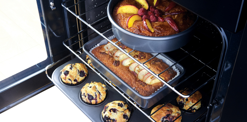 Ofen mit frisch gebackenen Muffins, Obstkuchen und Bananenbrot auf Backblech und Rost Offener Backofen mit einer runden Kuchenform, einem Bananenbrot und fünf Heidelbeermuffins beim Backen