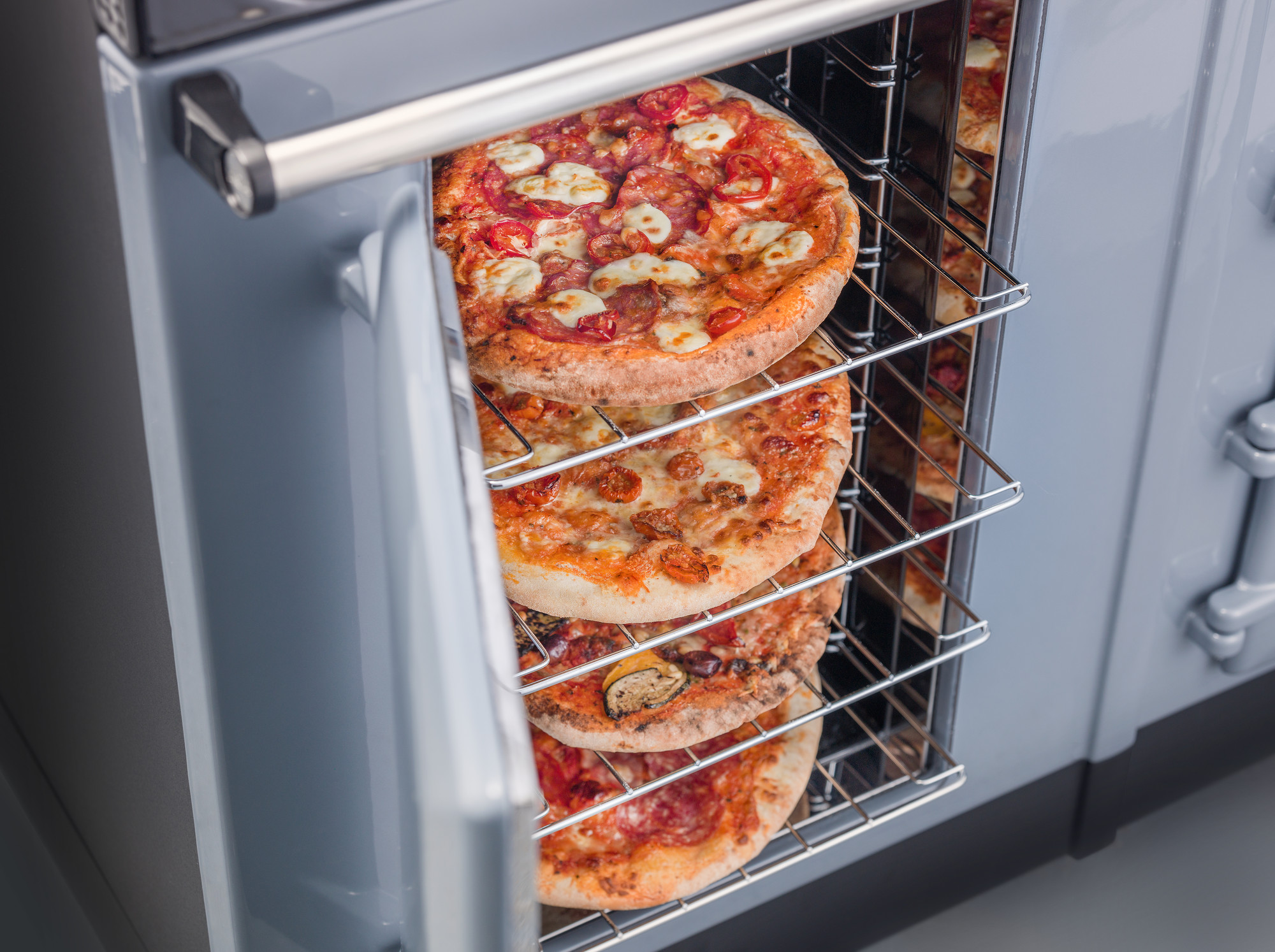 Vier frisch gebackene Pizzen im Ofen auf mehreren Rosten Mehrere belegte Pizzen werden gleichzeitig auf den Gitterrosten eines geöffneten Ofens gebacken