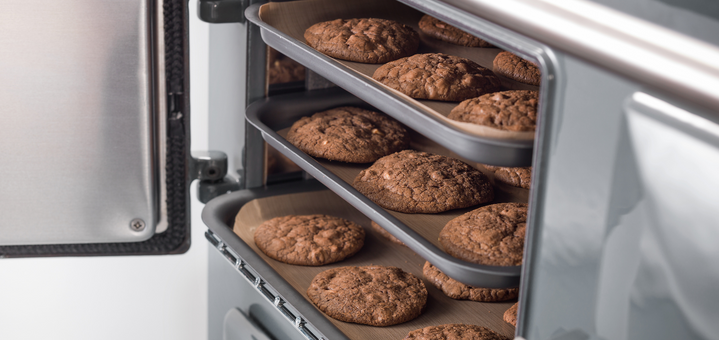 Frisch gebackene Schokoladenkekse auf Backblechen im Ofen Drei Backbleche mit Schokoladenkeksen werden in einem geöffneten Ofen gebacken.