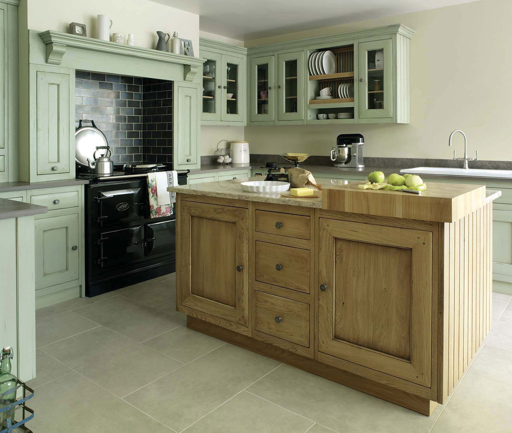 Landhausstil-Küche mit Kücheninsel aus Holz und grünen Schränken Helle Küche im Landhausstil mit großzügiger Kücheninsel aus Holz, grünen Küchenschränken, schwarzem Herd und Küchenaccessoires