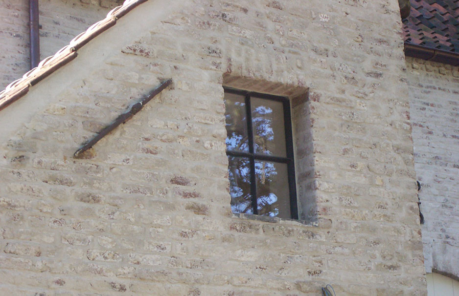 Historisches Fenster mit Sprossen in einer Natursteinfassade Quadratisches Fenster mit schwarzem Sprossenrahmen in einer Mauer aus Naturstein, spiegelt Bäume wider