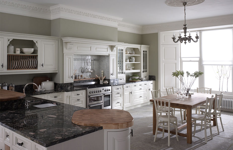 Moderne Landhausküche mit weißen Schränken, Granit-Arbeitsplatten und Essbereich Helle Landhausküche mit weißen Einbauschränken, dunklen Arbeitsplatten, großem Esstisch aus Holz und Kronleuchter am Fenster
