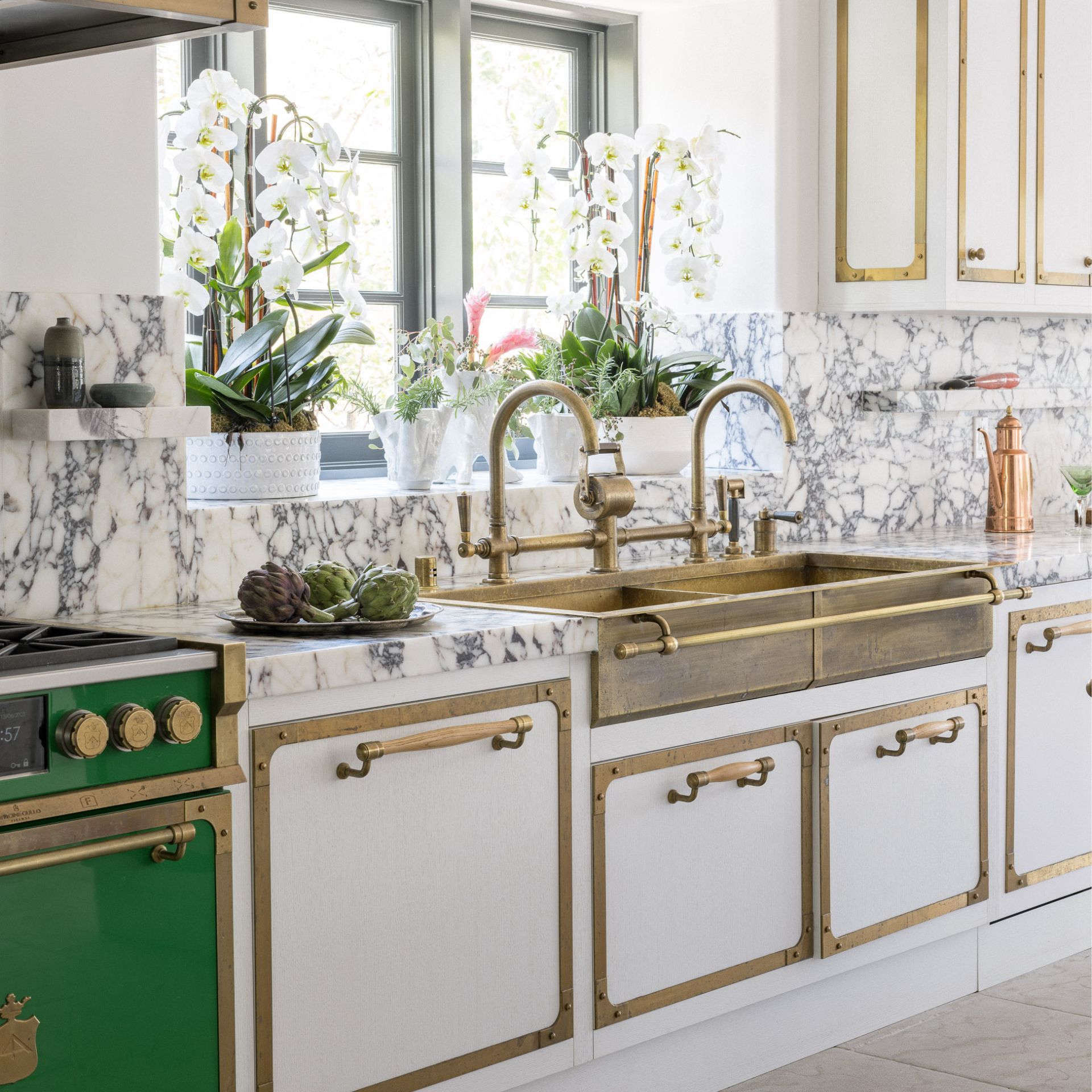 Hochwertige Messing-Küchenspüle mit Doppelarmatur, Marmor-Arbeitsplatte und weißen Fronten; stilvolle Küche mit Orchideen am Fenster – Produkt: Spülbecken und Armatur im Vintage-Look