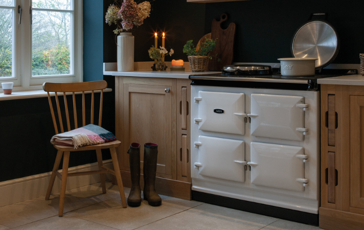 Gemütliche Landhausküche mit traditionellem weißen AGA-Herd, Holzstuhl, Gummistiefeln und frischen Blumen Helle Landhausküche mit AGA-Herd, Stuhl mit Decke, Gummistiefeln und dekorativen Blumenarrangements auf der Arbeitsfläche
