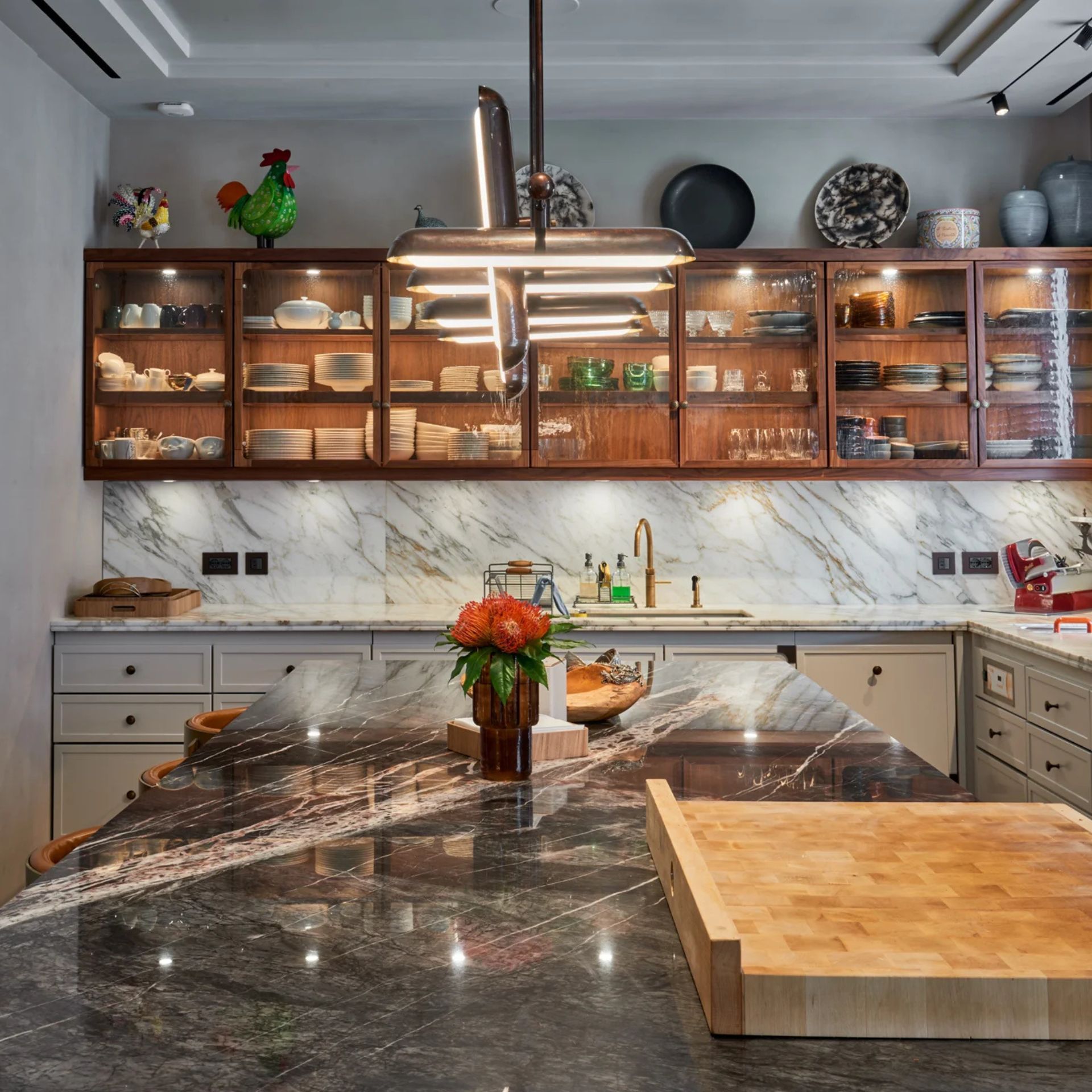 Kücheninsel mit dunkler Marmorarbeitsplatte, großem Holz-Schneidebrett, Glasoberschränken aus Holz, Messingarmatur und Design-Pendelleuchte – stilvolle Premium-Küche