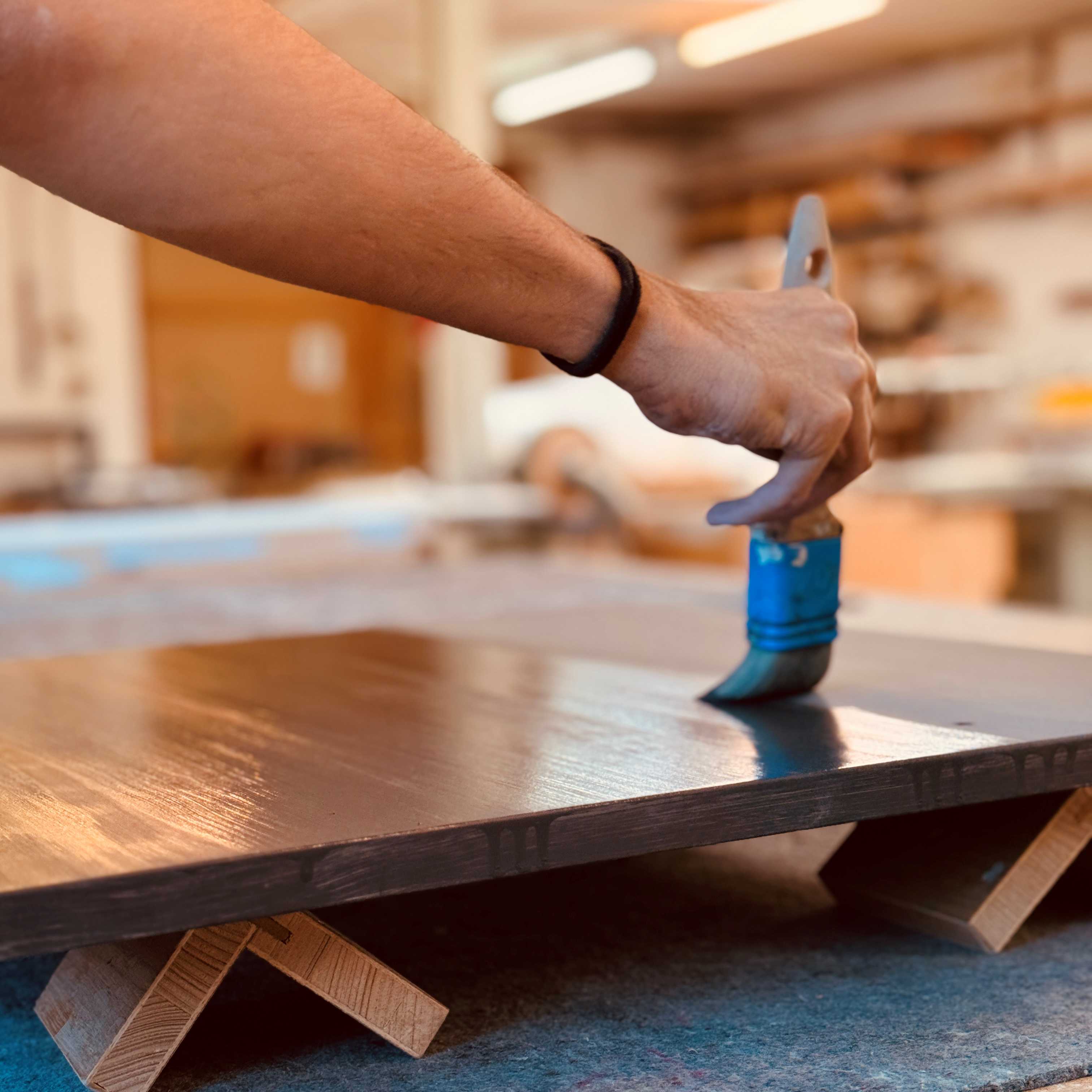 Hand trägt mit Pinsel eine dunkle Holzlasur auf eine aufgelegte Holzplatte auf – professionelle Oberflächenbehandlung und Versiegelung für Möbel
