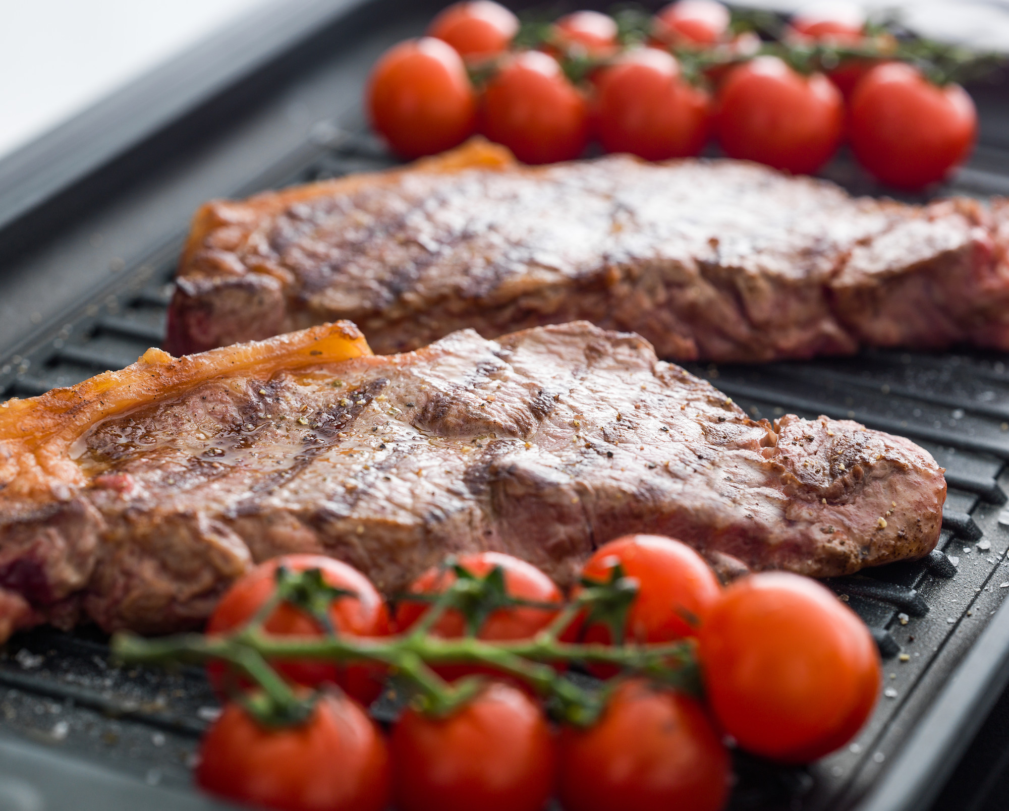 Saftige Rindersteaks und frische Kirschtomaten auf dem Grill - perfekt für Grillabende Zwei gegrillte Rindersteaks mit aromatischer Kruste und frischen Kirschtomaten auf einem Grillrost
