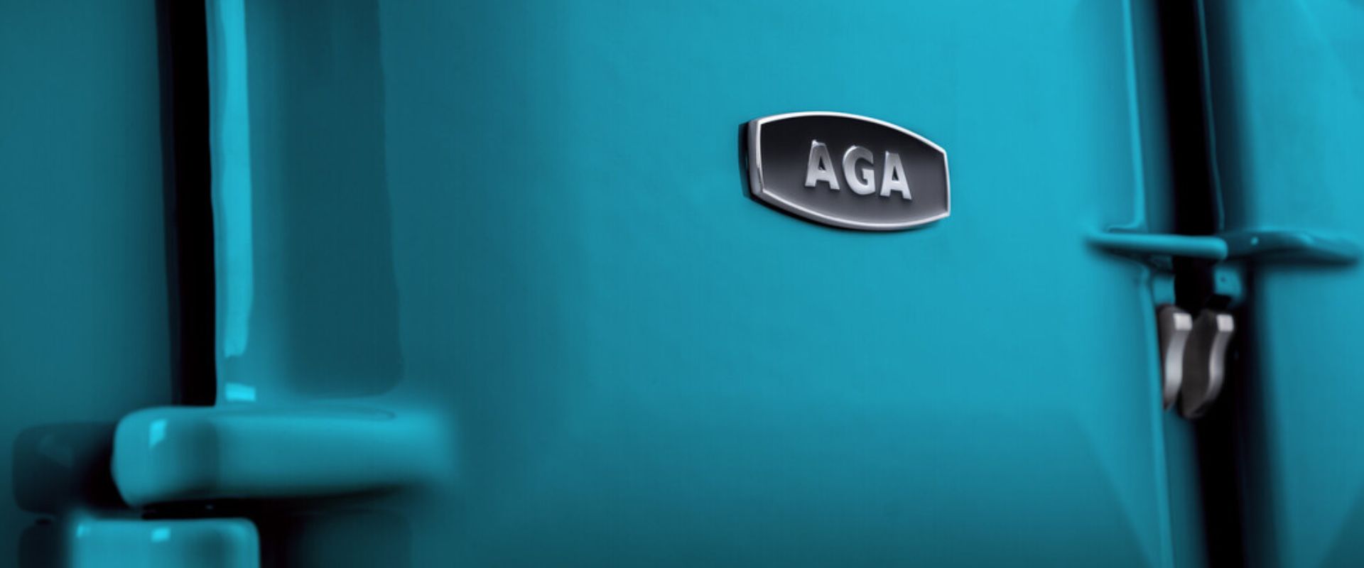 Nahaufnahme eines türkisfarbenen AGA Herdes: glänzende Emaillefront mit AGA-Logo und seitlichen Scharnier-/Griffelementen – Premium-Küchenofen Detail