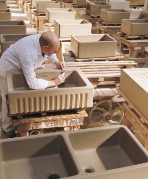 Handgefertigte Keramikspülen in der Produktion – Detailarbeit in der Manufaktur Ein Mitarbeiter bearbeitet sorgfältig eine Keramikspüle in einer Werkstatt, umgeben von weiteren Spülen auf Paletten.