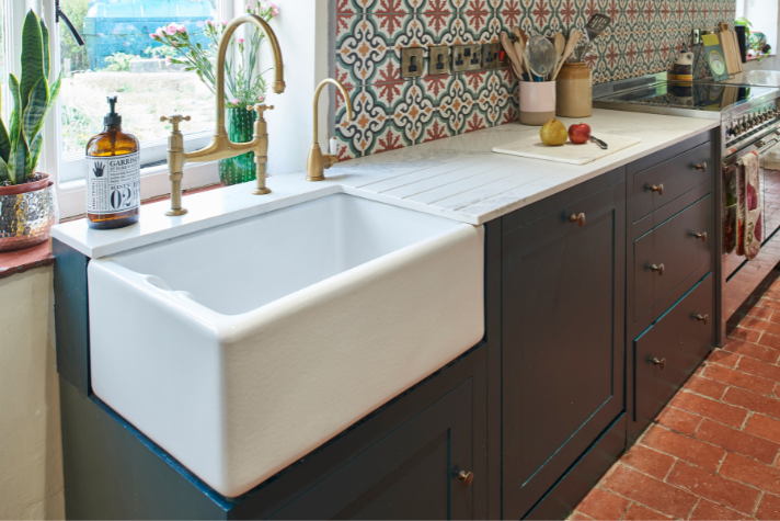 Landhausstil-Küche mit großer Keramik-Spüle, goldenen Wasserhähnen und gemusterten Fliesen Moderne Küche im Landhausstil mit weißer Keramikspüle, goldenen Armaturen, dunklen Unterschränken und gemusterten Wandfliesen neben einem Fenster