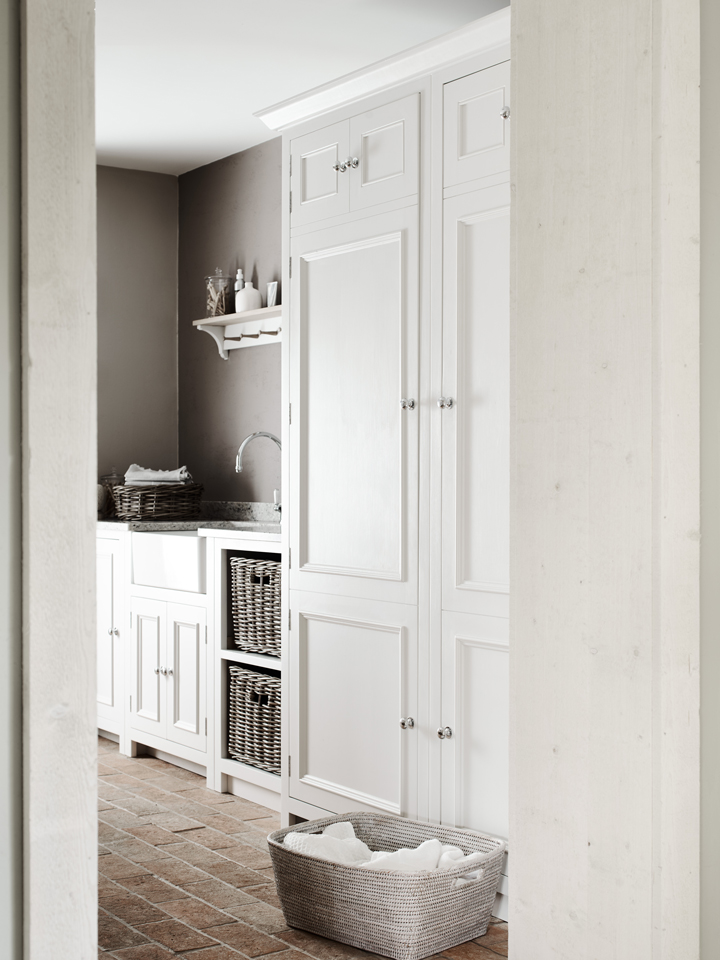 Helle, moderne Waschküche mit weißen Einbauschränken, Körben und gefliestem Boden Lichtdurchflutete Waschküche mit weißen Schränken, Körben aus Rattan, Waschbecken und gefliestem Boden