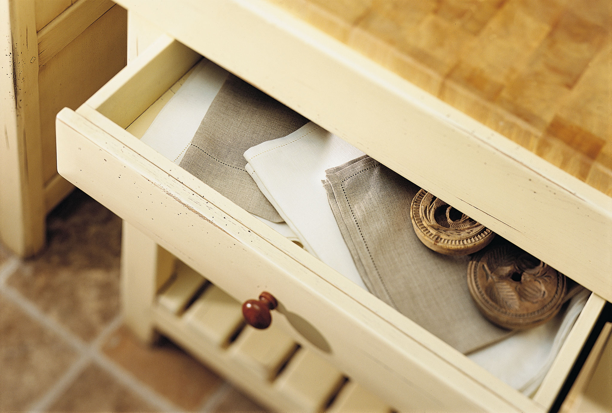 Offene Küchenschublade mit Leinenservietten und hölzernen Untersetzern auf heller Kommode Detailaufnahme einer offenen Schublade in einer cremefarbenen Küchenkommode, darin gefaltete Leinenservietten in Beige und Weiß sowie zwei hölzerne Untersetzer