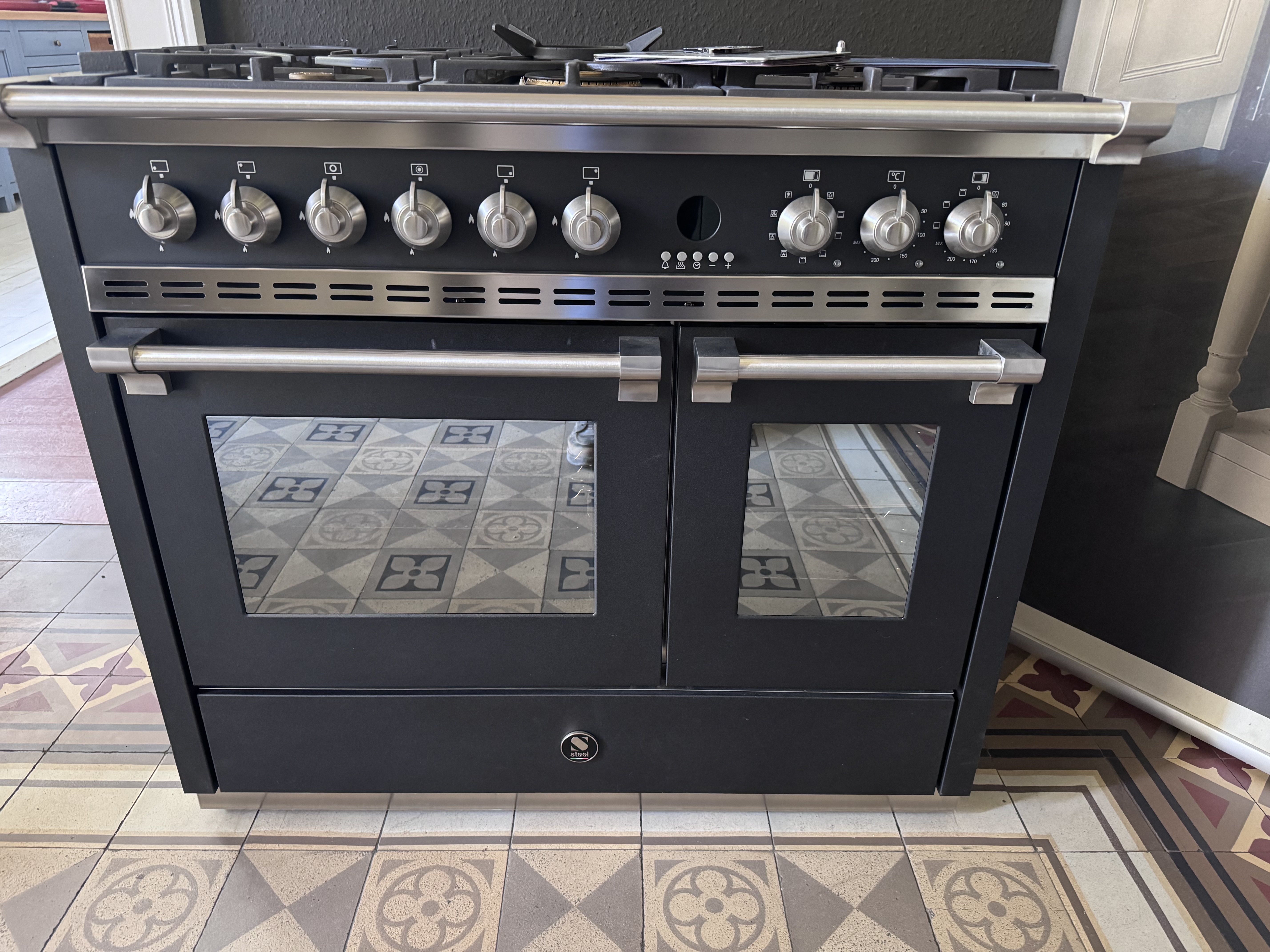 Schwarzer Standherd in Edelstahl-Optik mit Gaskochfeld, mehreren Drehknöpfen, Doppelbackofen und Glastüren; Frontansicht auf gefliestem Boden