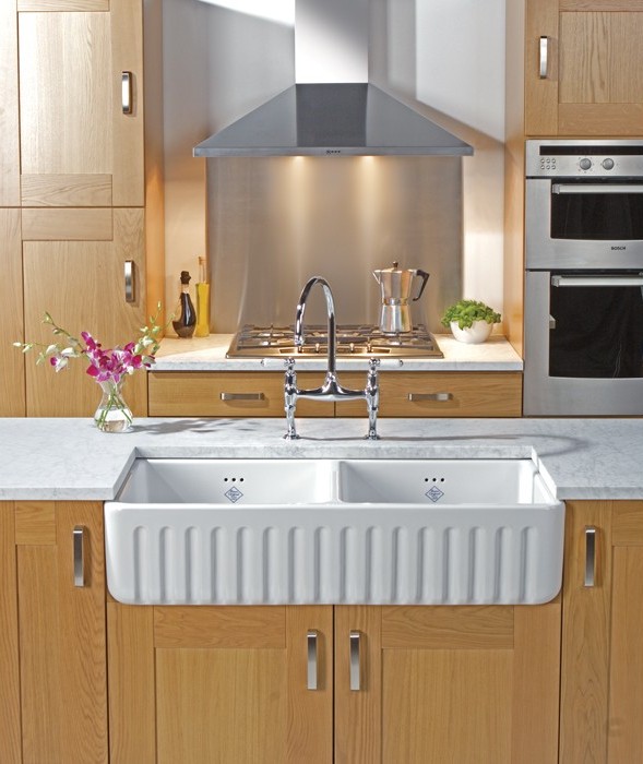 Moderne Küchenzeile mit Doppelspülbecken aus Keramik, Holzfronten und Edelstahl-Dunstabzugshaube Küchenzeile mit Doppelspülbecken aus weißer Keramik, Holzmöbeln, Edelstahl-Dunstabzugshaube und Herd, daneben Deko wie Blumen und Topfpflanze