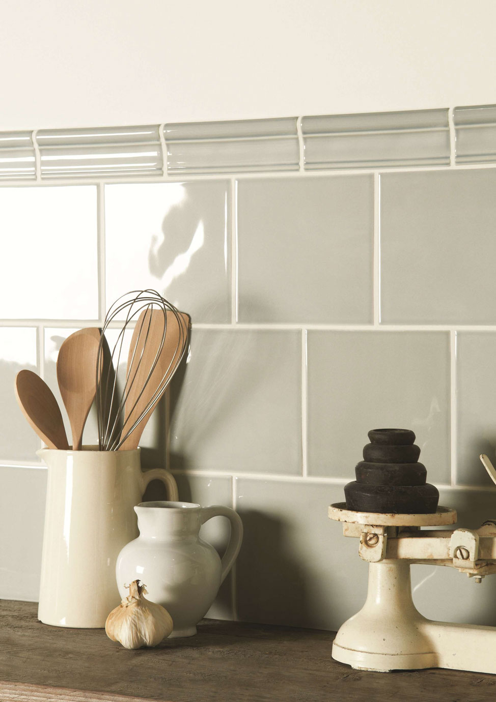 Küchenrückwand mit hellgrauen Metrofliesen im modernen Landhausstil Nahaufnahme einer Küchenarbeitsplatte mit cremefarbener Waage, Keramikkrug mit Holzkochlöffeln, Schneebesen, kleiner Milchkännchen und Knoblauch vor hellgrauen glänzenden Wandfliesen im Metrodesign