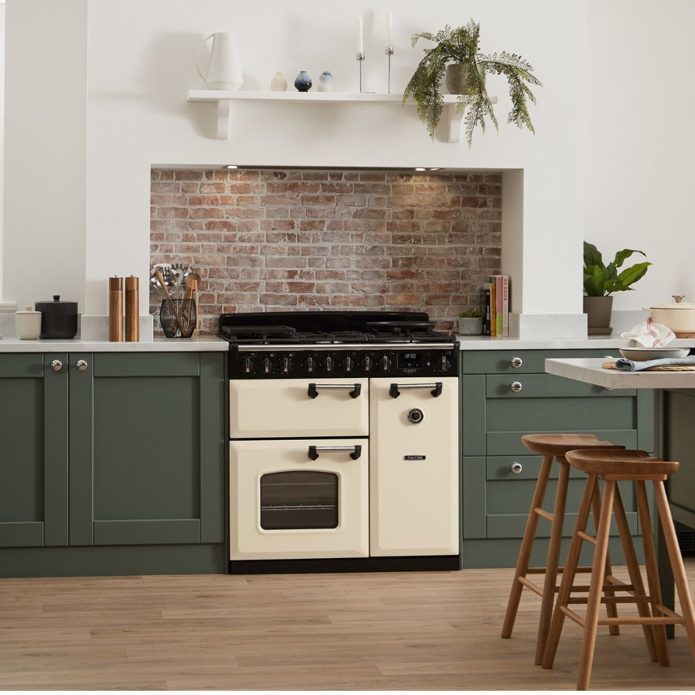 Freistehender cremefarbener Range Cooker mit Gas-Kochfeld, zwei Backöfen und Steuerknöpfen, integriert in grüne Shaker-Küche mit Backsteinrückwand