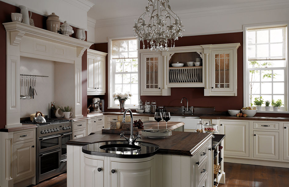 Elegante Landhausküche mit Kochinsel, cremefarbenen Schränken und Kronleuchter Gemütliche, klassische Landhausküche mit cremefarbenen Holzschränken, Kücheninsel, dunkler Arbeitsplatte, großer Herd, Kronleuchter und natürlichem Lichteinfall durch Fenster.