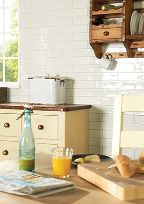 Gemütliche Küchenszene mit Frühstück, Weißbrot, Saft und Flasche auf Holztisch vor hellen Fliesen Frühstück auf Holztisch in heller Landhausküche mit Fliesenwand, Brot, Saft, Glasflasche und Küchenregal