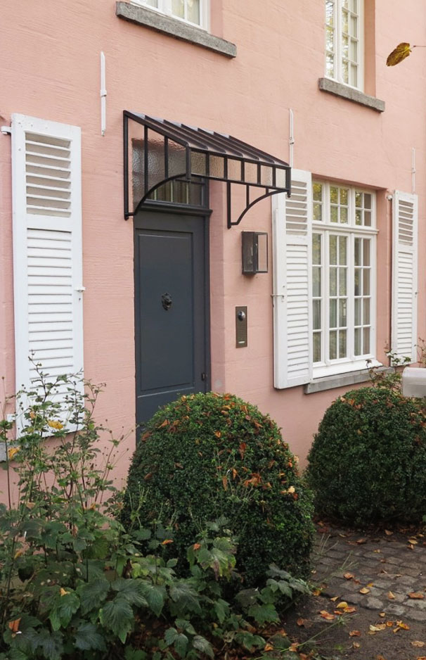 Eingangstür mit Vordach und weißen Fensterläden an rosa Hausfassade Schwarze Haustür mit Glaskuppel-Vordach, daneben weiße Fensterläden und große grüne Buchskugeln vor einem rosa Haus