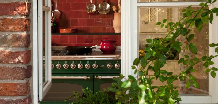 Eleganter grüner Standherd (Range Cooker) mit mehreren Backöfen und Drehknöpfen, rote Fliesenrückwand, Töpfe und Pfanne – Blick durchs offene Fenster mit Pflanzen