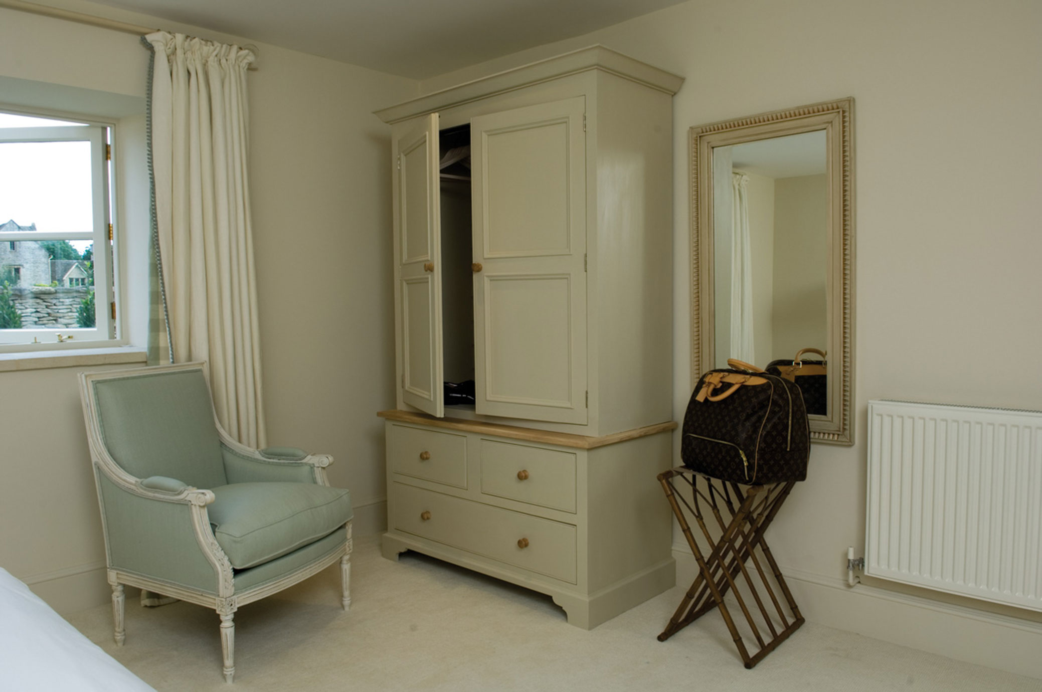 Helles Schlafzimmer mit Sessel, Kleiderschrank, Spiegel und Reisetasche Schlafzimmer mit hellgrünem Sessel, cremefarbenem Kleiderschrank, großem Wandspiegel und einer Reisetasche auf einem Gepäckständer