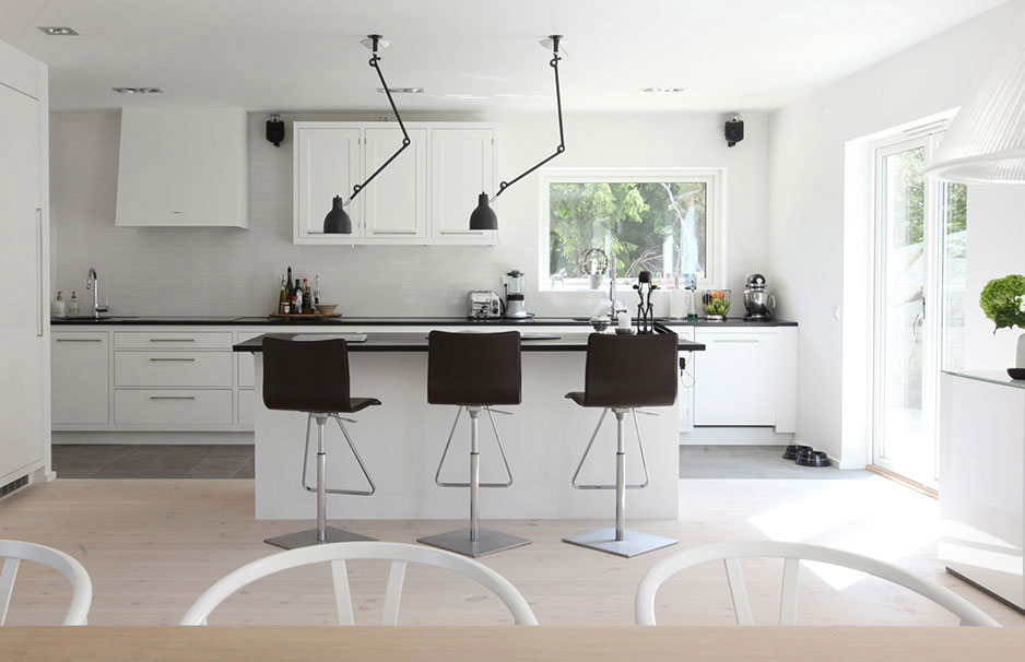 Moderne weiße Küche mit Kochinsel, Hockern und großen Fenstern für viel Tageslicht Helle, moderne Einbauküche mit weißer Kochinsel, drei dunklen Barhockern und großen Fenstern, die viel natürliches Licht hereinlassen