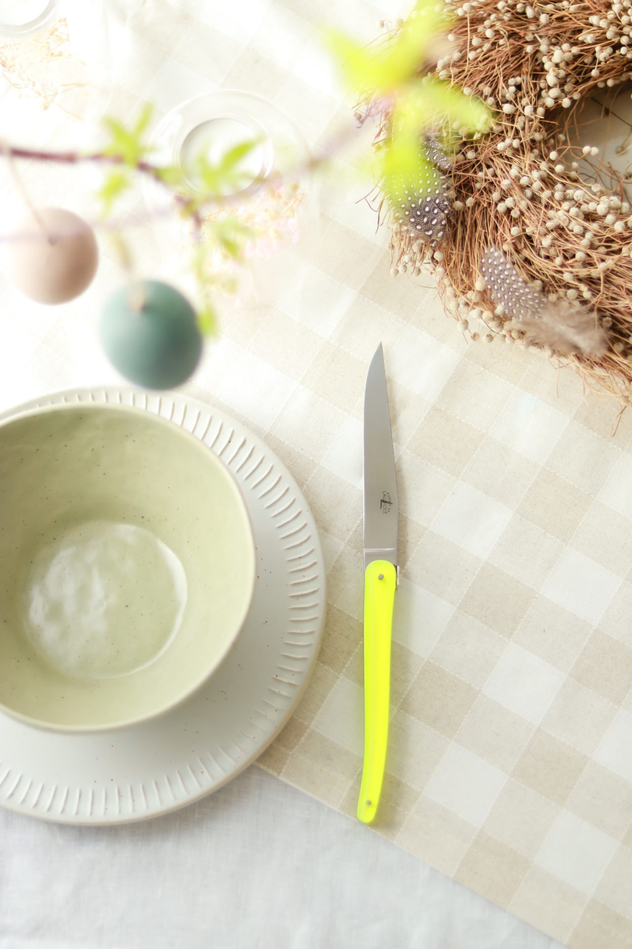Frühlinghafte Tischdekoration mit gelbem Messer, Keramikteller, Schale und Osterkranz Helles, österliches Tischarrangement mit gelbem Messer, Keramikschale, Unterteller und dekorativem Kranz auf hell karierter Tischdecke