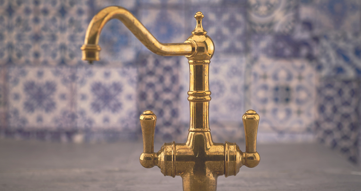 Antiker Wasserhahn aus Messing mit klassischem Design vor verzierter Fliesenwand Nahaufnahme eines nostalgischen, goldfarbenen Wasserhahns im Vintage-Stil vor einer Wand mit blauen Ornamentfliesen