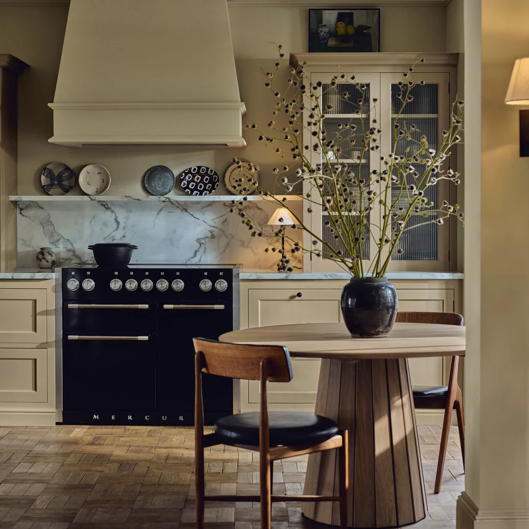 Runder Esstisch aus Massivholz mit zwei Stühlen; Vase mit Zweigen auf dem Tisch, dahinter beige Landhausküche mit Marmor-Rückwand und schwarzem Range Cooker
